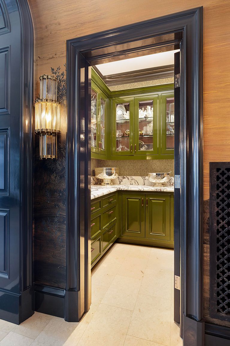 This image showcases a well-organized pantry with olive green cabinetry, marble countertops, and glass-fronted upper cabinets displaying dishware and glassware. The pantry is accessed through a dark-framed doorway, adding a touch of elegance and sophistication. The space is illuminated by recessed lighting, creating a warm and inviting atmosphere.