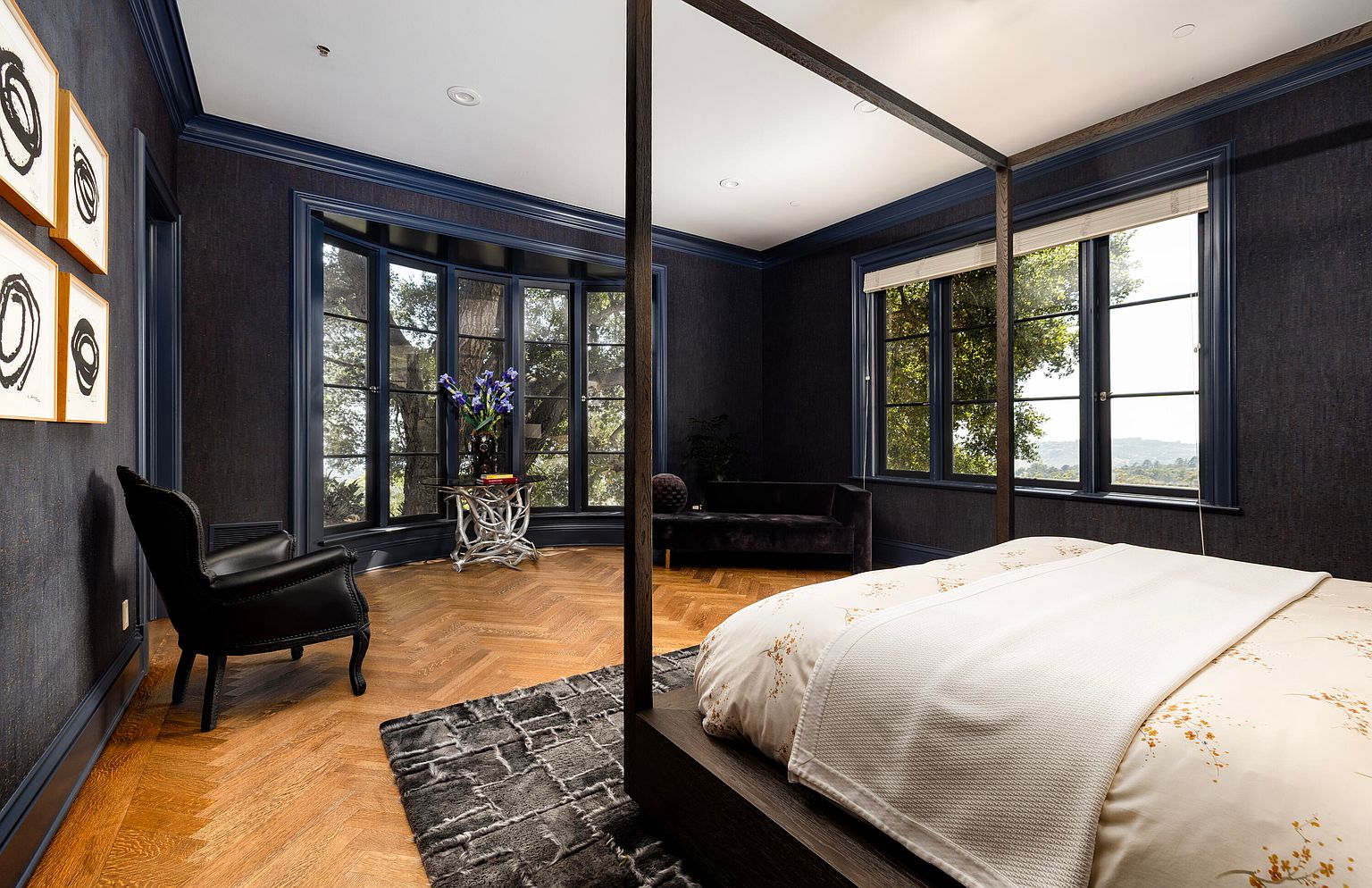 This is a luxurious primary bedroom featuring dark blue walls and trim, a four-poster bed with white linens, and a herringbone wood floor. A bay window offers natural light and views of the surrounding trees, while a seating area with a black chair and a dark velvet sofa provides additional comfort. The room exudes a sophisticated and elegant ambiance.