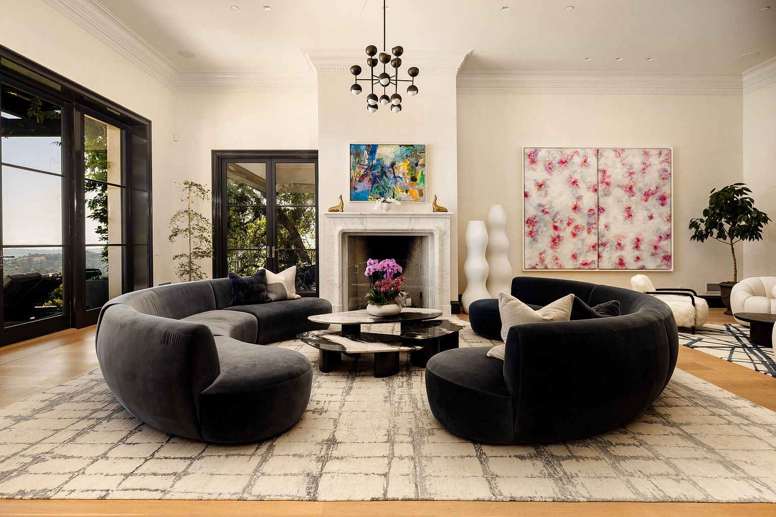 This is a spacious living room featuring a modern design with curved sofas arranged around a unique multi-tiered coffee table. The room is well-lit, with natural light streaming in through large windows and doors, complemented by a striking chandelier above the fireplace. The neutral color palette and artistic decor create an elegant and inviting atmosphere.