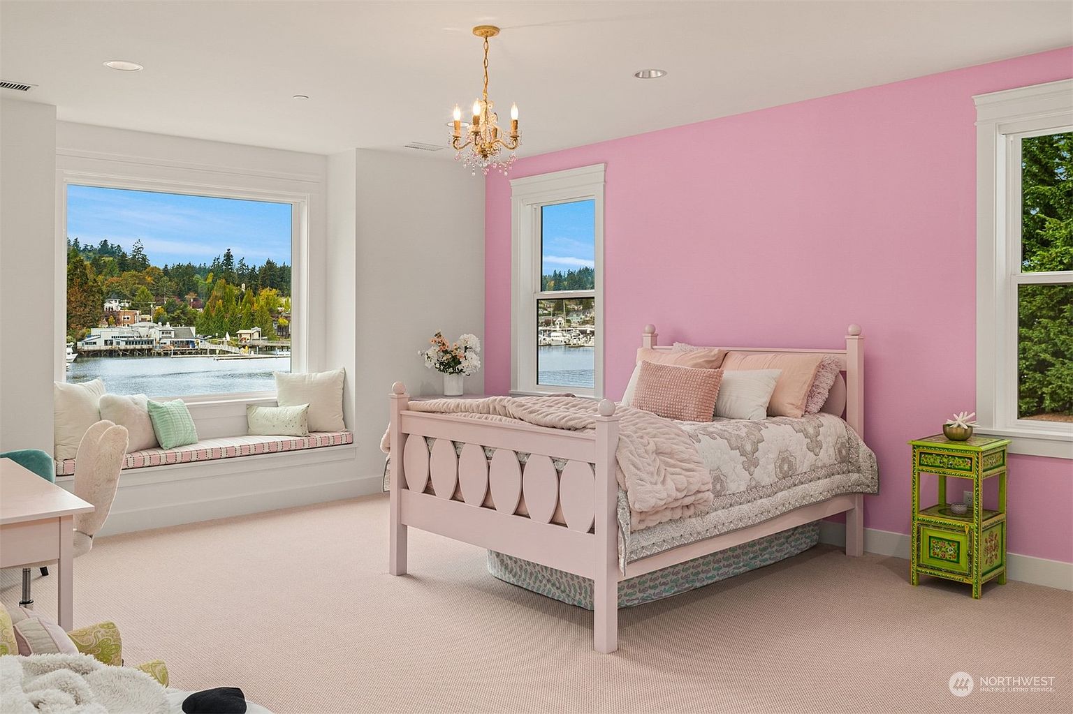 This bright and airy bedroom features a soft pink color palette, highlighted by a prominent pink accent wall and matching bed frame. A large window seat offers a picturesque view of the waterfront, while a crystal chandelier adds a touch of elegance to the space. The room is completed by a vibrant green bedside table and plush carpeting, creating a cheerful and inviting atmosphere.