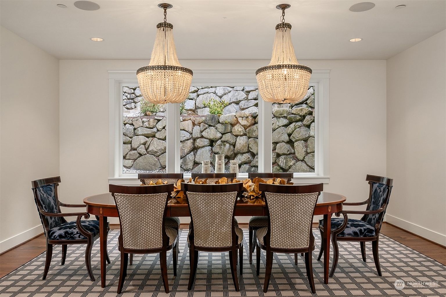 This formal dining room features a large, dark-toned oval table surrounded by six chairs with a mix of patterned fabric and woven backrests. Two ornate, beaded chandeliers hang symmetrically above the table, which is centered in front of a large window looking out onto a natural stone retaining wall. The room is grounded by a geometric patterned rug, creating a sophisticated and balanced atmosphere.