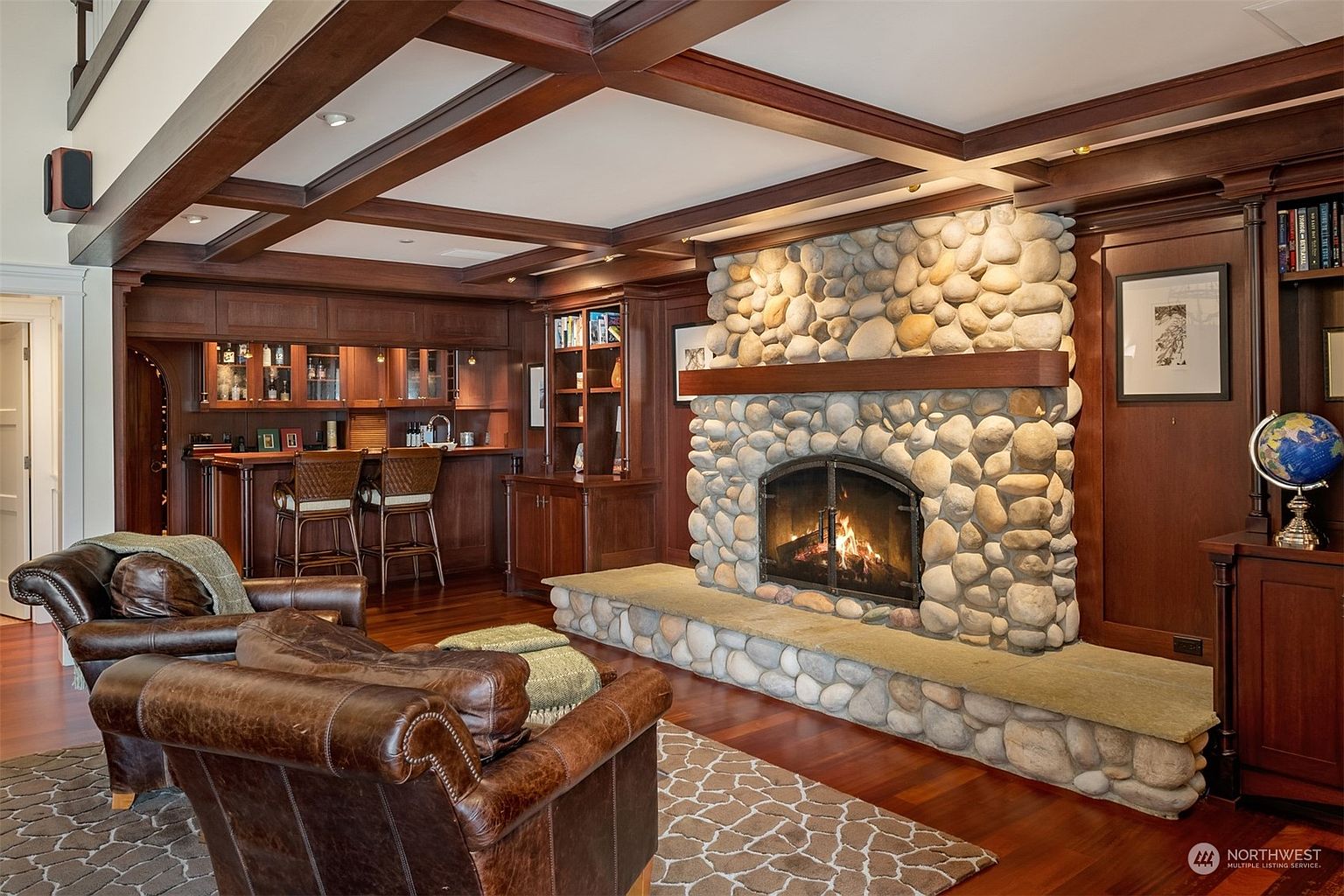 This sophisticated living room features a prominent stone-clad chimney breast with a wood mantel and a built-in fireplace, serving as the focal point of the space. The room is accented by rich wood paneling, a coffered ceiling, and a wet bar area with barstool seating, creating a warm and inviting atmosphere. A pair of leather armchairs sits on a patterned rug, providing a comfortable seating arrangement in this classic, study-like environment.