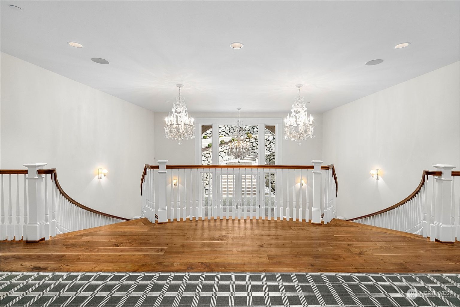 This grand foyer features a symmetrical, sweeping staircase with white balusters and a rich wood handrail that leads down to a lower level. The space is illuminated by two ornate crystal chandeliers and wall sconces, creating an elegant and formal atmosphere. A patterned runner adds texture to the landing, which overlooks a glass-paneled entryway that provides a view of the exterior stone landscaping.