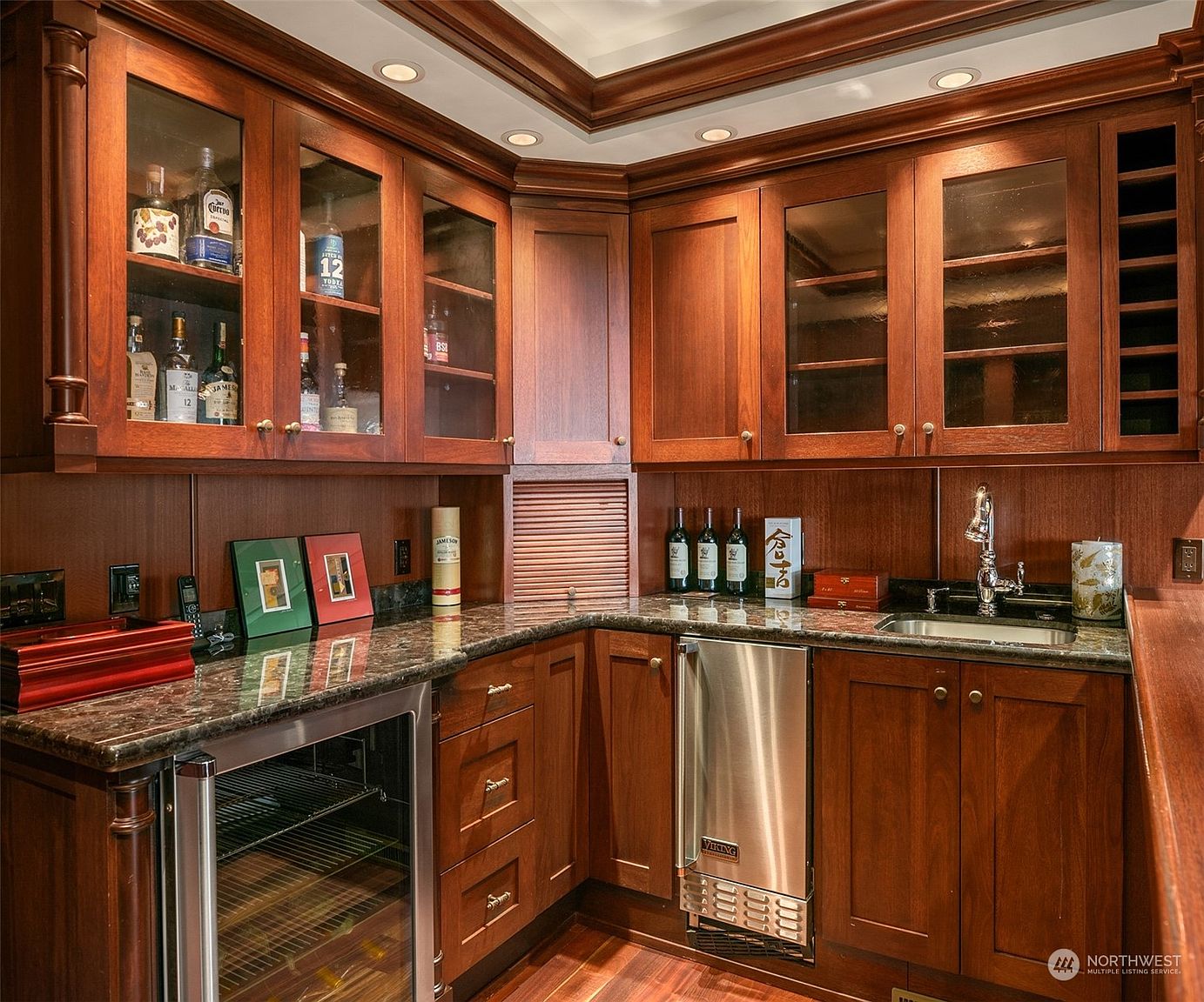 This elegant wet bar area features rich, dark-toned wood cabinetry with glass-front upper cabinets and integrated lighting. The space is equipped with a stainless steel under-counter refrigerator, a built-in ice maker, and a small sink set into a dark stone countertop. The warm, sophisticated aesthetic is enhanced by recessed ceiling lights and a dedicated wine rack, creating a refined atmosphere for entertaining.