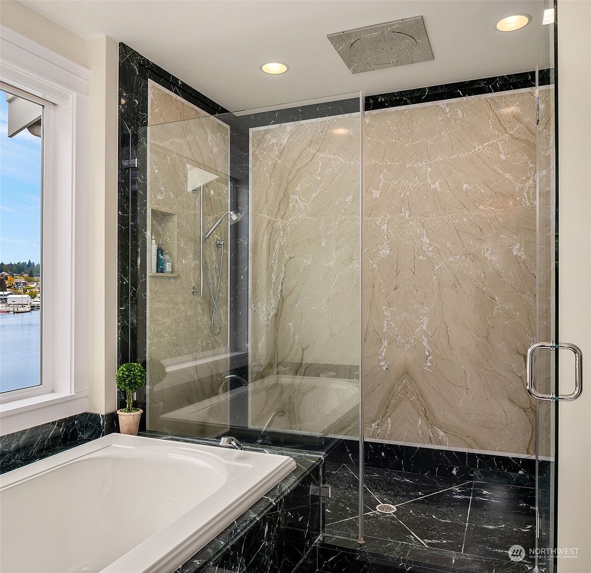 This luxurious primary bathroom features a large soaking tub set against a dark stone surround, adjacent to a spacious walk-in shower enclosed by glass panels. The shower interior is finished with light-toned, veined stone slabs and accented with dark stone trim, creating a sophisticated contrast. A window beside the tub provides a scenic view of the water, enhancing the serene and high-end atmosphere of the space.