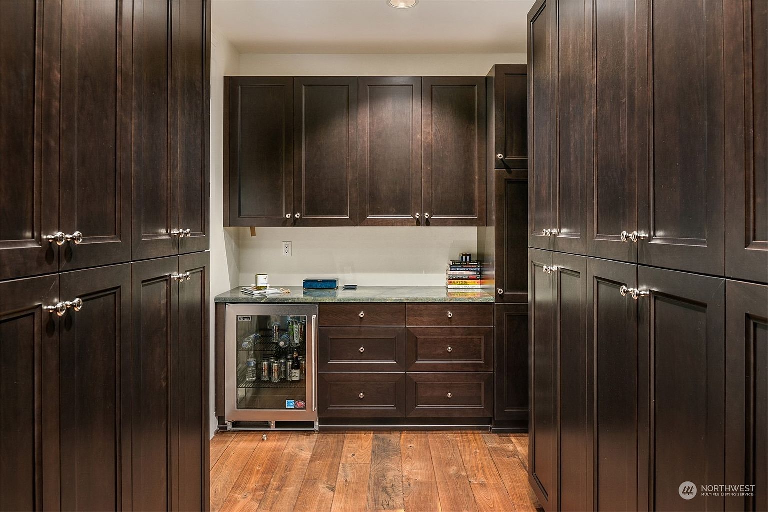 This butler's pantry or wet bar area features rich, dark wood cabinetry and a stone countertop, creating a sophisticated and moody aesthetic. The space is flanked by tall, matching storage cabinets on both sides and includes a built-in under-counter beverage refrigerator. The warm-toned wood flooring adds a natural element to the refined, enclosed design.