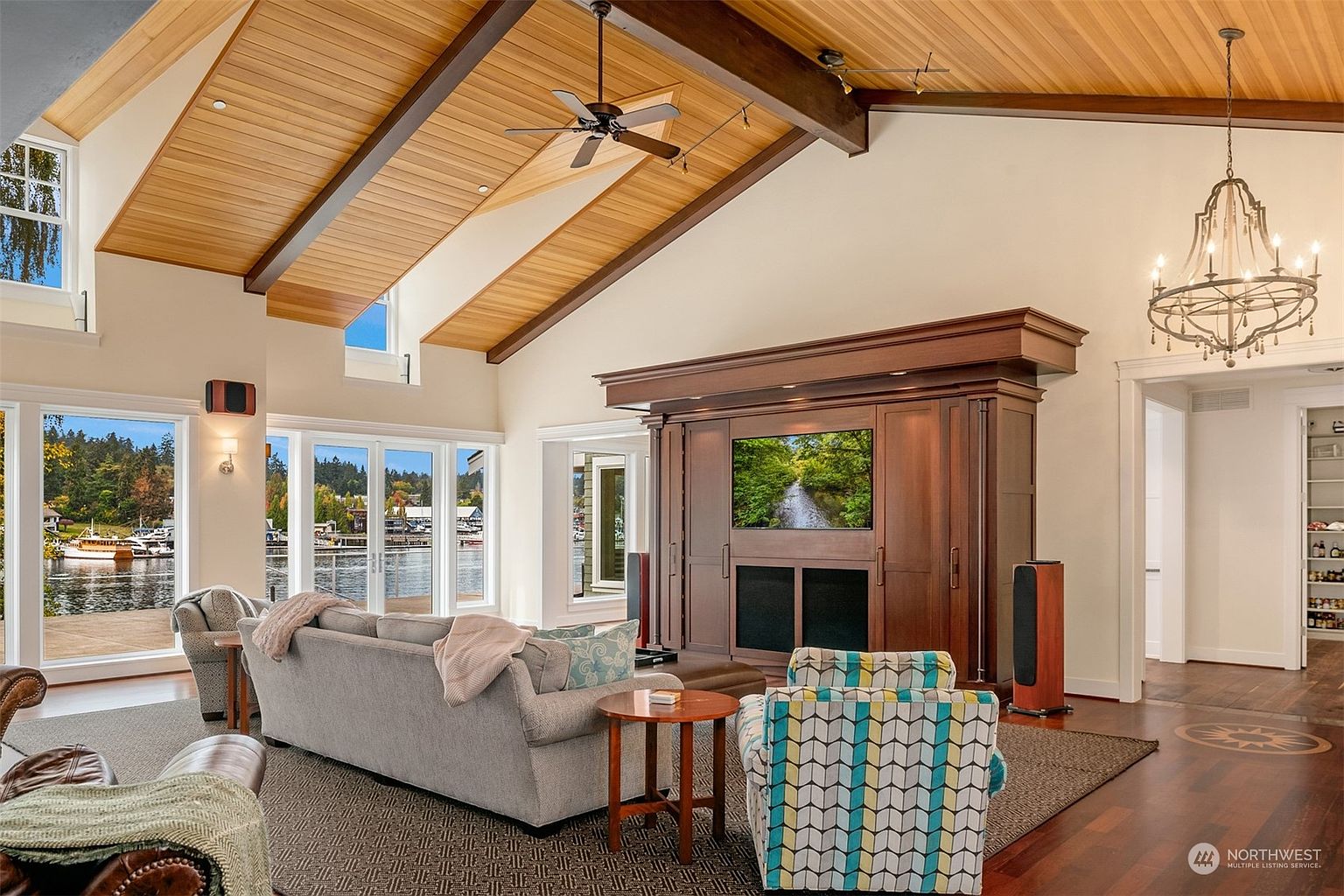 This spacious living room features a dramatic vaulted ceiling with exposed wood beams and warm wood paneling. A large, dark wood entertainment center serves as a focal point, flanked by tall speakers, while a comfortable seating area is arranged on a textured rug. Large windows and glass doors provide an expansive view of the waterfront, filling the room with natural light and creating an airy, inviting atmosphere.