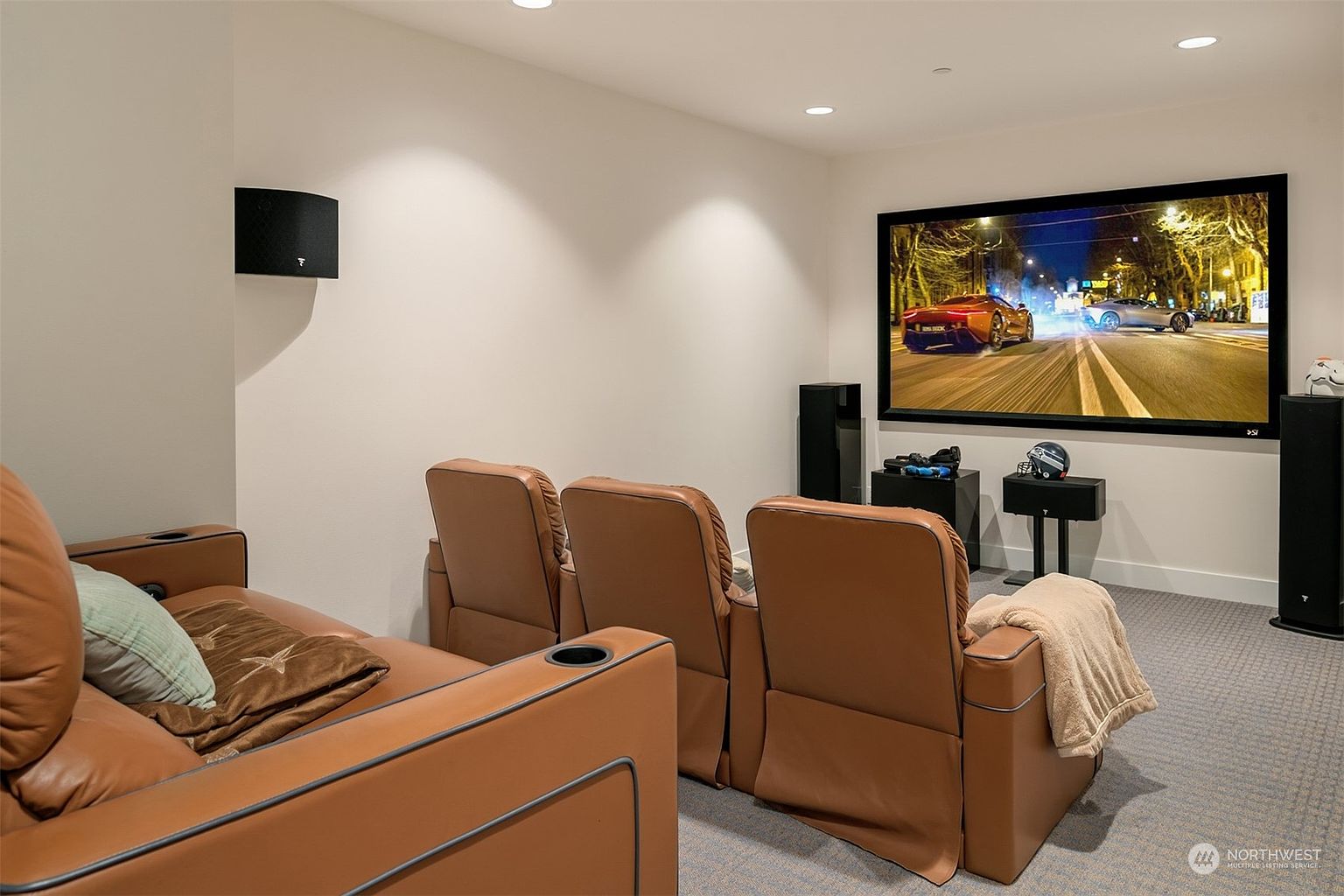 This dedicated home theater room features a large projection screen displaying a high-speed car chase, complemented by rows of comfortable, tan leather reclining seats. The space is designed for immersive entertainment, with wall-mounted speakers and a minimalist aesthetic that keeps the focus on the screen. The room is carpeted and illuminated by recessed ceiling lights, creating a cozy and private cinematic environment.