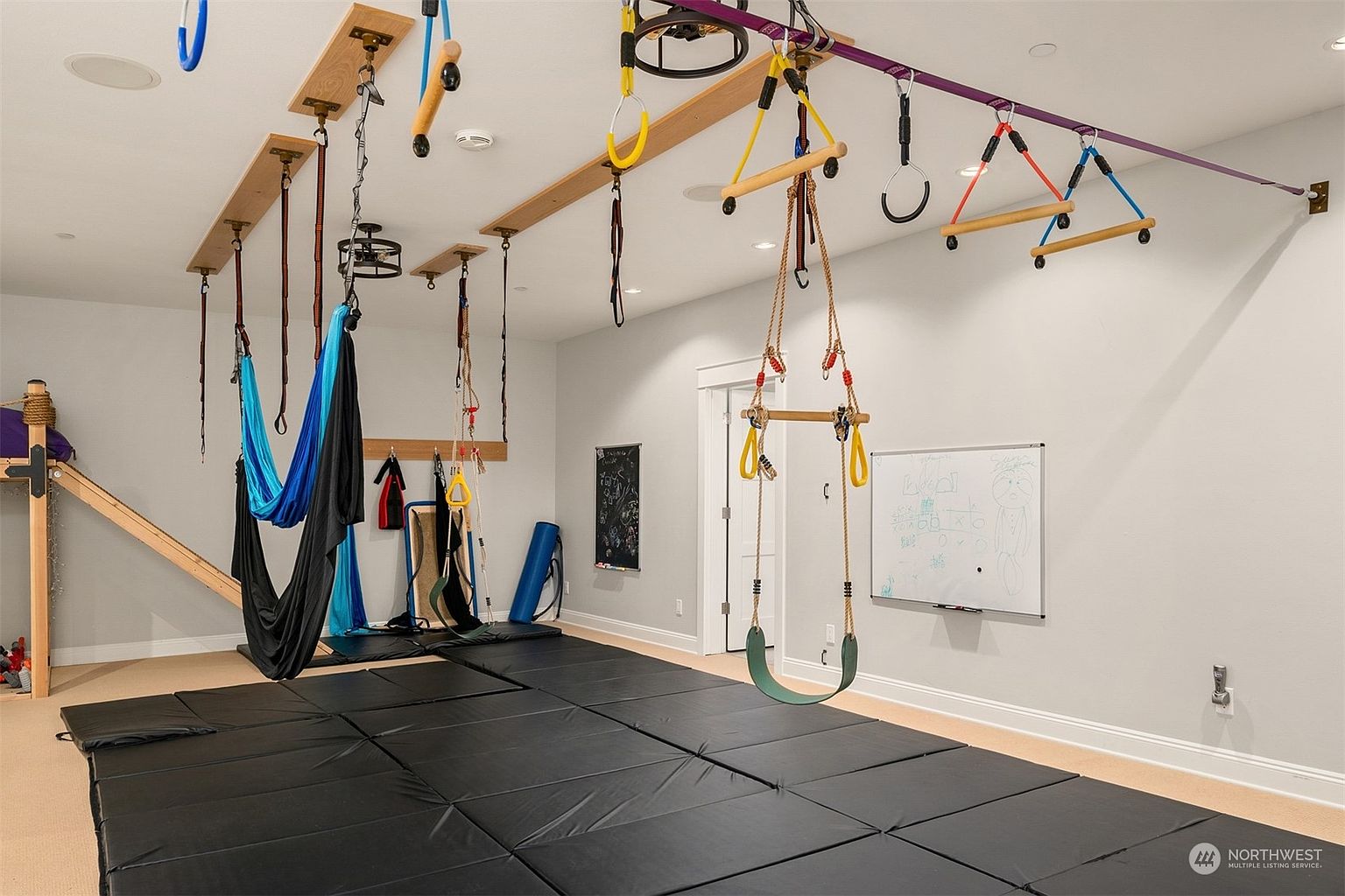 This dedicated indoor play and exercise room features a variety of suspended equipment, including swings, aerial silks, and climbing rings attached to ceiling-mounted wooden beams. The floor is covered with large, black protective mats, creating a safe environment for physical activity. A whiteboard and a chalkboard are mounted on the light-colored walls, adding functional elements to this versatile recreational space.