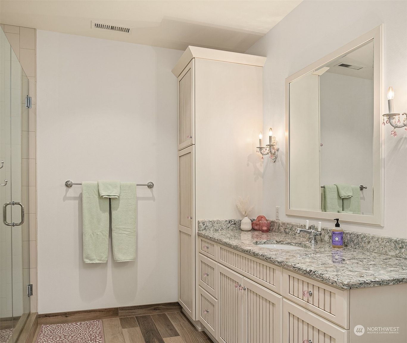 This bright bathroom features a vanity with light-colored, vertical-paneled cabinetry topped with a speckled stone countertop and an integrated sink. A tall, matching storage cabinet stands beside the vanity, while a large mirror is mounted above, flanked by decorative wall sconces. To the left, a glass-enclosed shower stall and a towel rack mounted on the wall complete the functional layout.