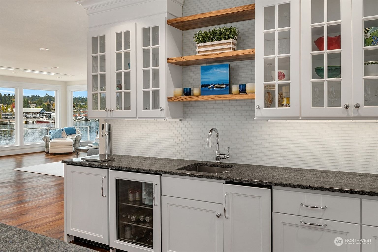 This elegant wet bar area features white cabinetry, a dark stone countertop, and a small under-counter beverage refrigerator. Open wood shelving is mounted against a light-colored subway tile backsplash, while a stainless steel sink and faucet complete the functional space. The perspective looks past the bar into an adjacent living area with large windows that provide a scenic view of the water.