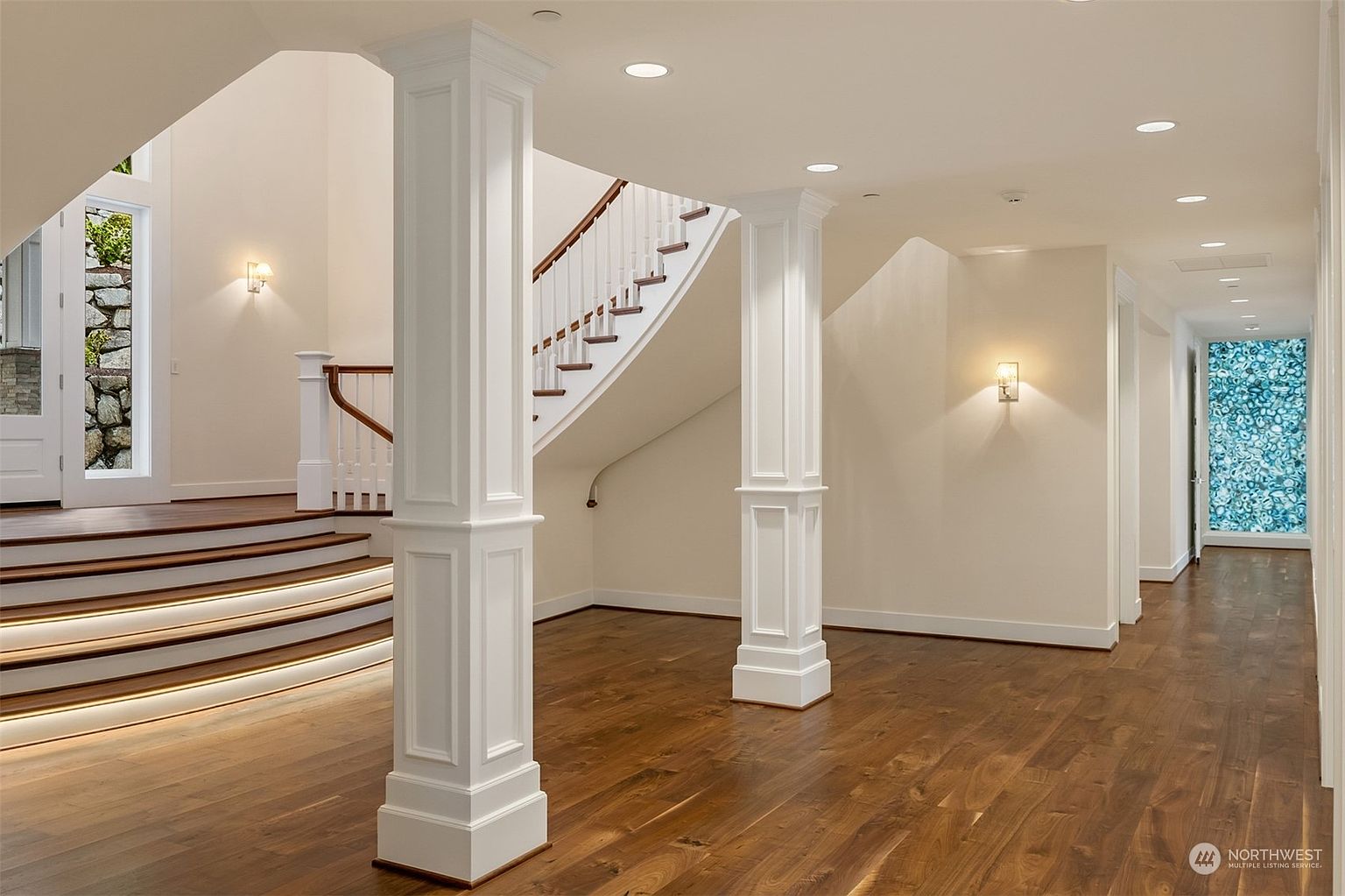 This elegant foyer features a grand curved staircase with a dark wood handrail and white balusters, complemented by warm wood flooring. Two prominent white columns frame the transition between the entryway and the hallway, while recessed lighting and wall sconces provide soft illumination. A striking, backlit blue decorative panel adds a unique artistic focal point at the end of the hallway, creating a sophisticated and welcoming atmosphere.