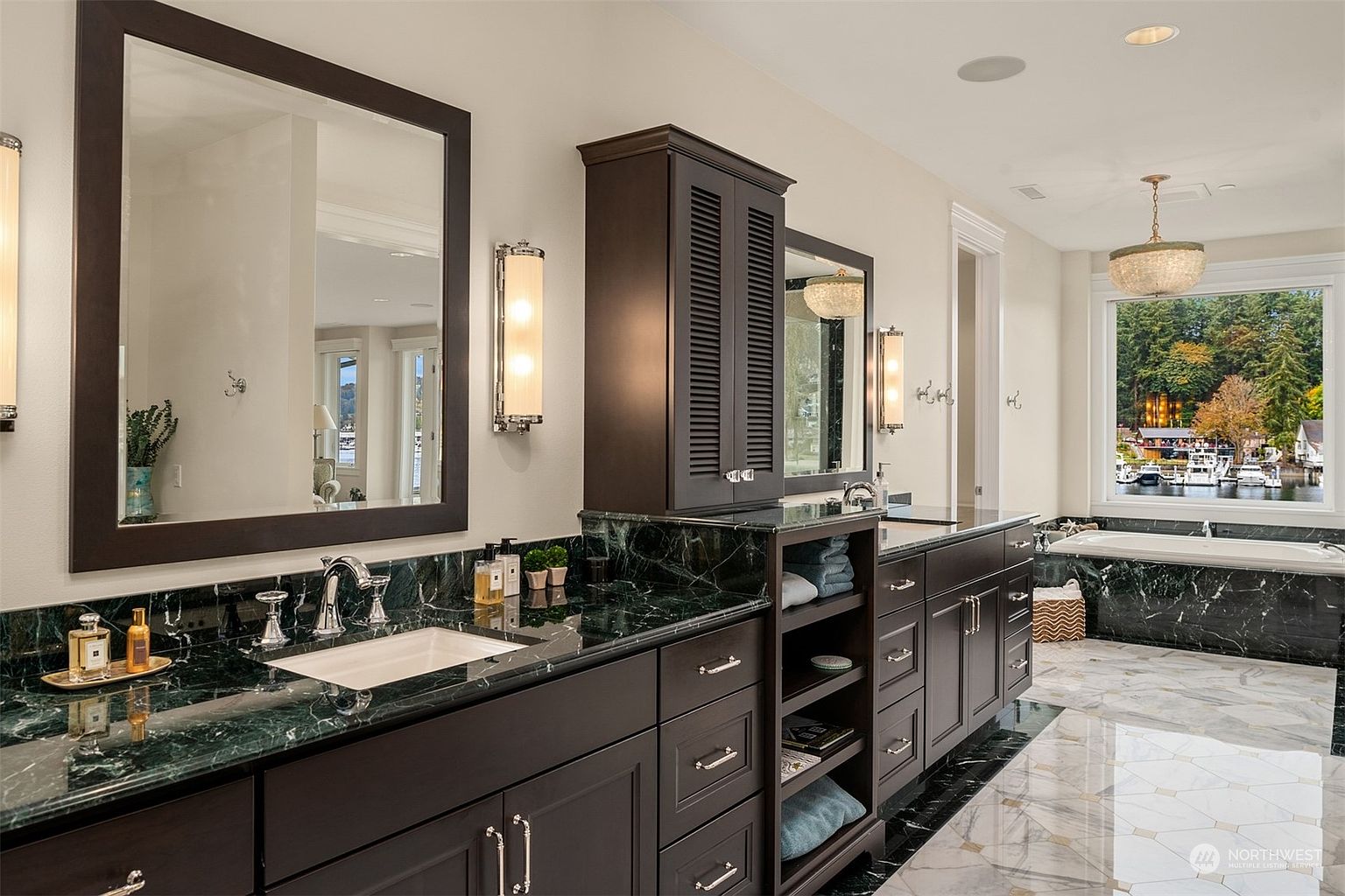 This luxurious primary bathroom features a long vanity with dark cabinetry and a striking green stone countertop. The space is accented by large mirrors, elegant wall sconces, and a deep soaking tub positioned near a large window with a scenic water view. The room is finished with polished patterned tile flooring, creating a sophisticated and high-end atmosphere.