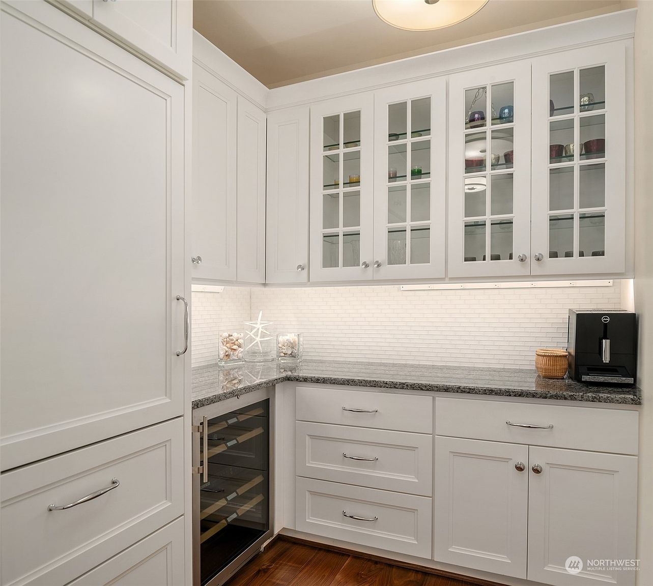 This well-appointed kitchen wet bar features crisp white cabinetry with glass-front upper doors and a dark, speckled stone countertop. The space includes a built-in wine refrigerator, a coffee station, and a clean white tile backsplash illuminated by under-cabinet lighting. The perspective is a straight-on view of the cabinetry arrangement, highlighting the organized storage and elegant finishes.