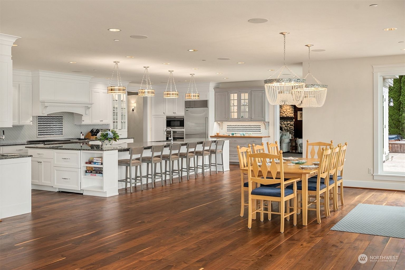 This spacious, open-concept kitchen features a large central island with bar seating, white cabinetry, and dark stone countertops. A dining area with a wooden table and chairs sits adjacent to the kitchen, illuminated by a prominent crystal chandelier. The room is anchored by rich, dark wood flooring and recessed lighting, creating a bright and inviting atmosphere with a view into an adjacent living space.