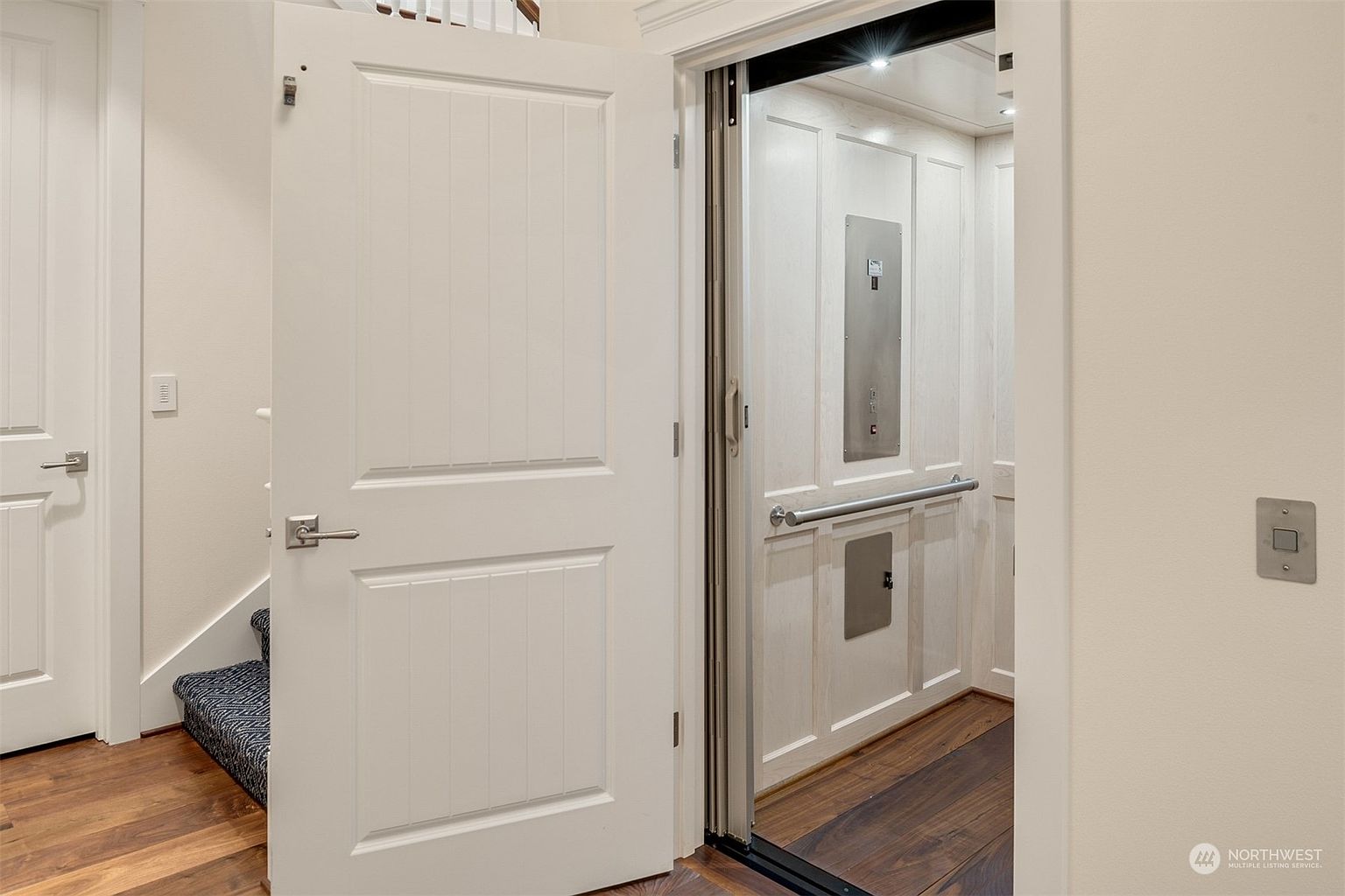 This interior view captures a residential elevator with a paneled interior and a metal handrail, positioned next to a white door leading to a staircase. The flooring consists of dark wood planks, while the adjacent staircase features a patterned runner. The perspective is eye-level, highlighting the accessibility features and the clean, neutral aesthetic of the home's transition space.