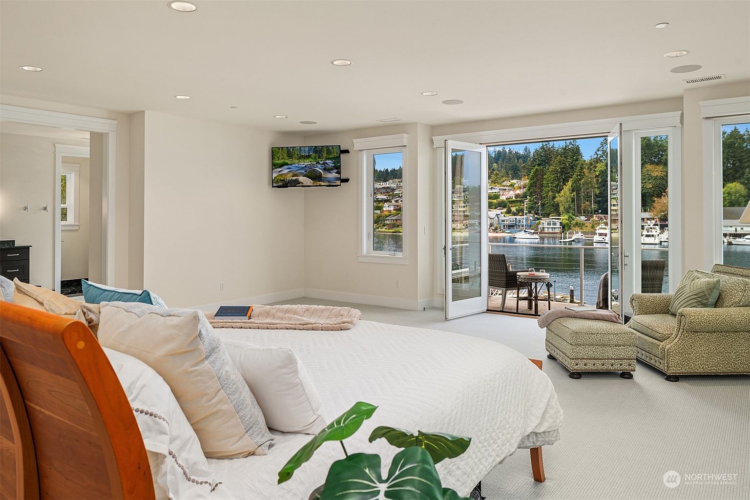 This spacious primary bedroom features a large bed with a wooden headboard and a cozy seating area with an armchair and ottoman. The room opens through double glass doors onto a private balcony that offers a scenic view of the water and surrounding landscape. The neutral color palette and soft lighting create a serene atmosphere, while a wall-mounted television provides entertainment.