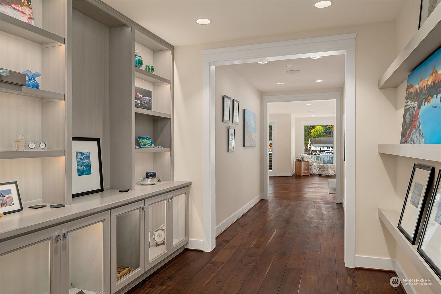 This elegant hallway features rich, dark wood flooring and light-toned walls, creating a sophisticated transition between living spaces. To the left, a custom built-in shelving unit with integrated lighting displays decorative items, while the right side features floating shelves adorned with framed artwork. The perspective draws the eye through a wide, cased opening toward a bright, sunlit room with a view of the water beyond.