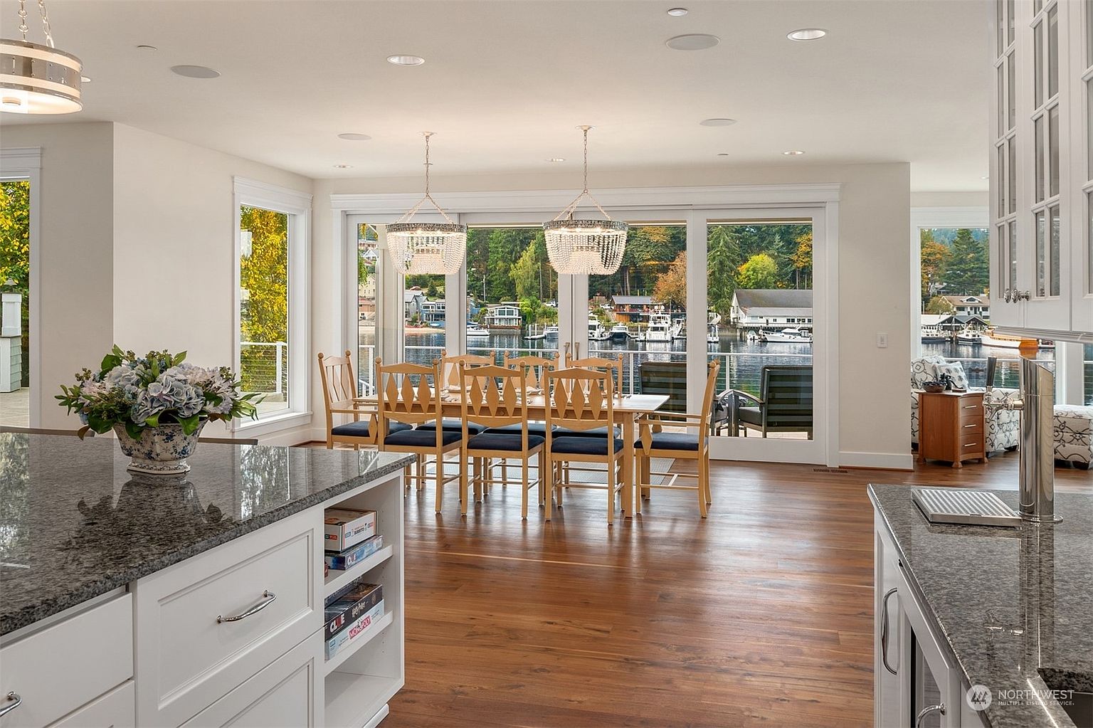 This bright, open-concept kitchen features a large central island with a dark stone countertop and white cabinetry, seamlessly transitioning into a dining area. The space is anchored by a dining table set for eight, positioned in front of expansive glass doors that offer a panoramic view of the water and surrounding trees. Two elegant beaded chandeliers hang above the dining table, while the warm wood flooring and neutral color palette create an inviting and sophisticated atmosphere.