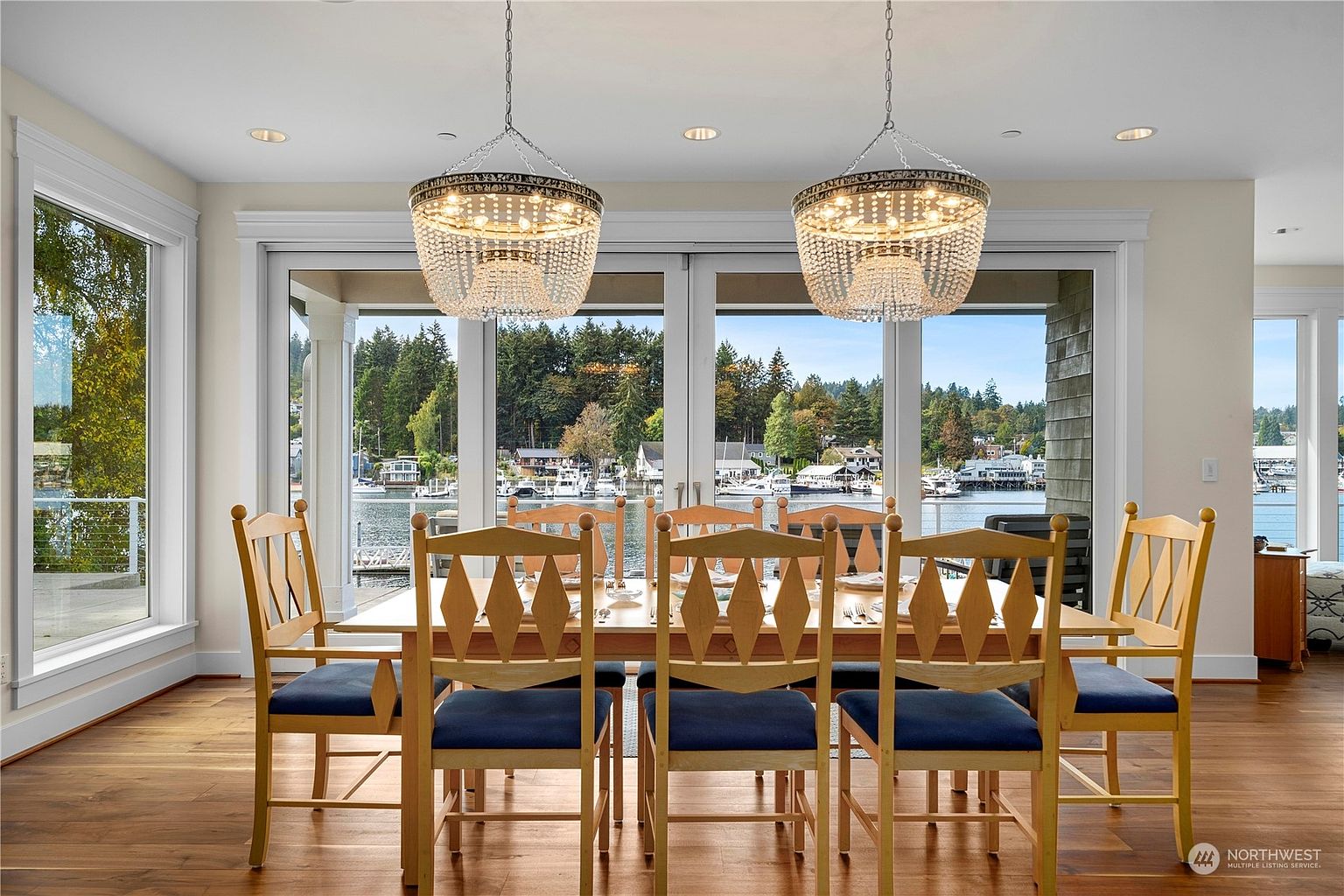This elegant dining room features a light wood table surrounded by eight chairs with dark blue upholstered seats. Two ornate, crystal-adorned chandeliers hang above the table, serving as a focal point. Large glass doors provide a panoramic view of the waterfront, creating a bright and airy atmosphere with a seamless connection to the outdoors.