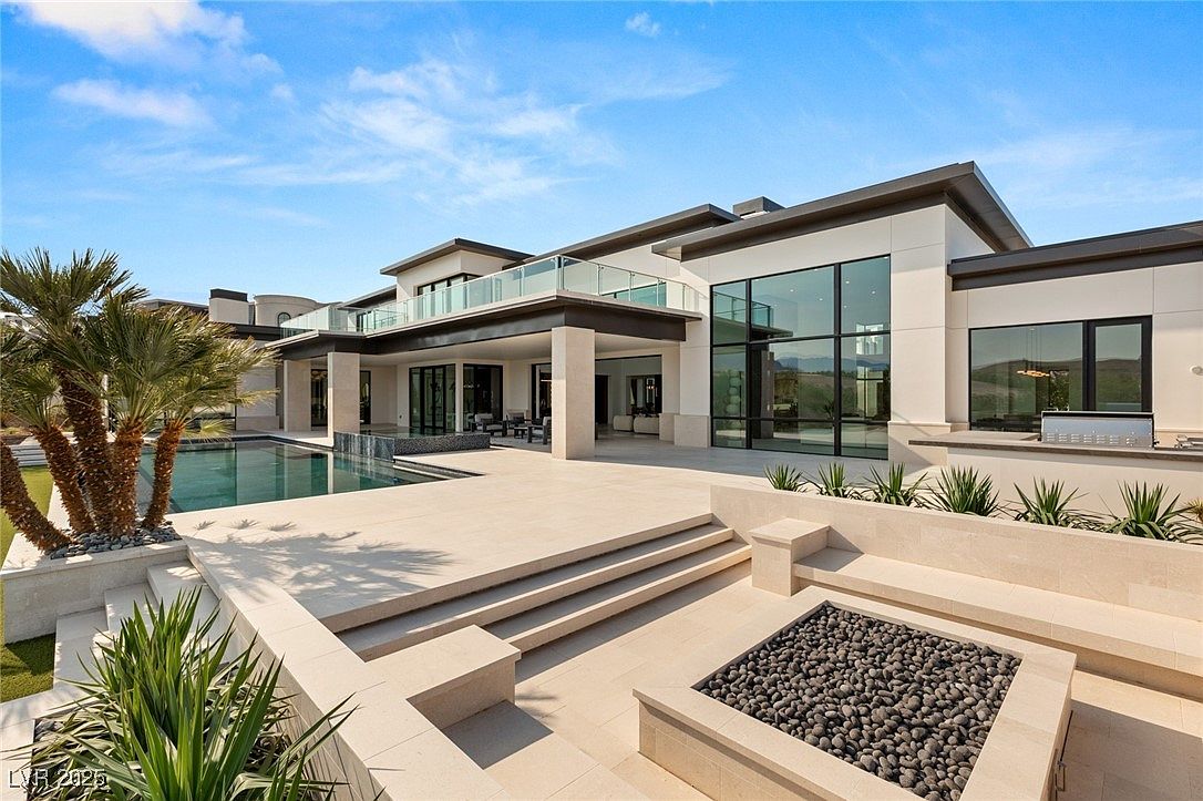 This is a rear exterior view of a modern luxury home, showcasing a large patio area with a fire pit, a swimming pool, and well-manicured landscaping. The architecture features clean lines, large windows, and a balcony, creating an inviting outdoor living space perfect for entertaining and relaxation. The perspective is from a low angle, emphasizing the scale and grandeur of the property.