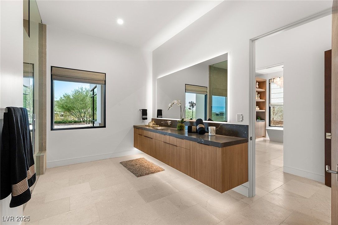 This is a modern primary bathroom featuring a floating vanity with a dark countertop and wood-paneled cabinets. A large, illuminated mirror spans the length of the vanity, and a window provides natural light and a view of the outdoors. The flooring is light-colored tile, and the overall aesthetic is clean and minimalist.
