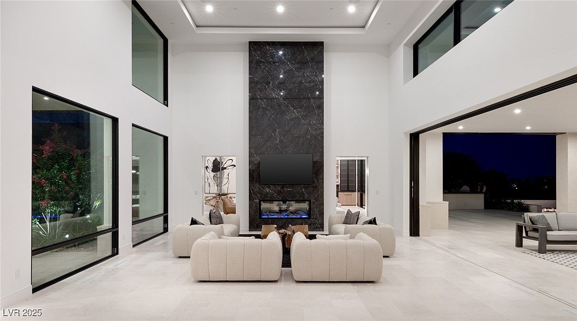 This is a spacious living room featuring high ceilings and large windows that provide ample natural light. The focal point is a modern fireplace with a dark marble surround and a mounted television. The room is furnished with contemporary sofas arranged around a central coffee table, creating an inviting and luxurious atmosphere.