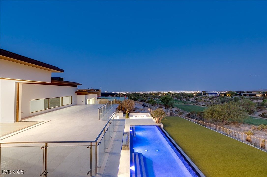 This image showcases the rear exterior of a modern luxury home at dusk, featuring a sleek infinity pool with illuminated water, a well-manicured lawn, and a spacious patio area with glass railings. The house boasts clean architectural lines and large windows, offering stunning views of the surrounding landscape and city lights in the distance. The perspective is from an elevated position, providing a comprehensive view of the property's outdoor living spaces.