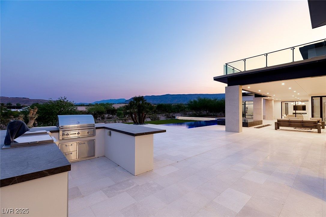 This image showcases a luxurious outdoor living space with a modern design. The patio features a built-in outdoor kitchen with a stainless steel grill, a sleek infinity pool, and a covered lounge area with comfortable seating. The balcony above offers additional space and views of the surrounding landscape, creating an inviting and sophisticated atmosphere.