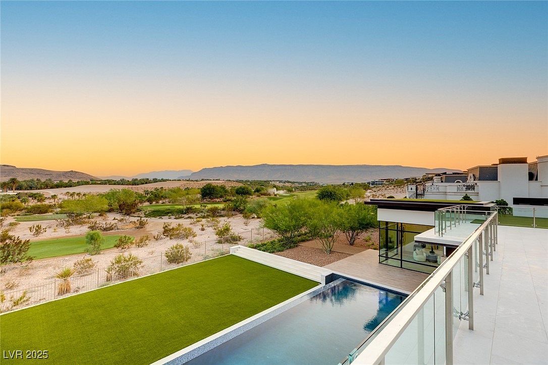 This image showcases a luxurious outdoor pool area with a modern aesthetic. The pool is surrounded by a well-manicured lawn and a sleek deck, complemented by a glass railing that offers unobstructed views of the landscape. The property overlooks a scenic vista with mountains in the distance, creating a serene and upscale ambiance.