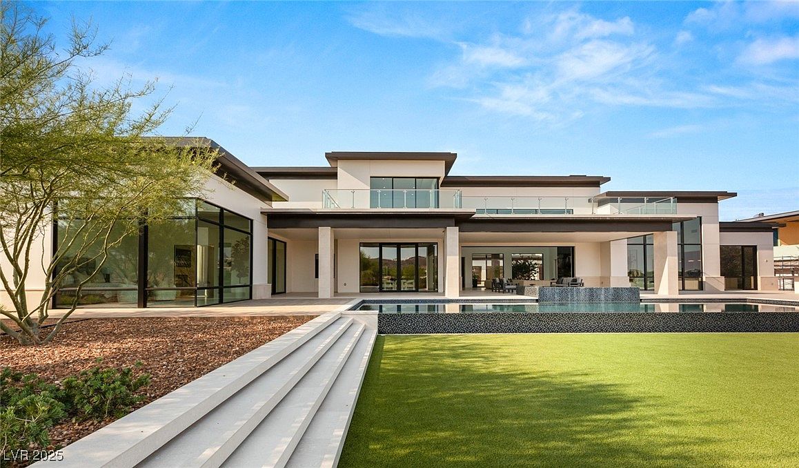 This image showcases the rear exterior of a modern luxury home, featuring a sleek design with clean lines and a flat roof. The property includes a swimming pool with a unique water feature, a well-manicured lawn, and a patio area perfect for outdoor entertaining. Large glass windows and doors provide ample natural light and seamless indoor-outdoor living.