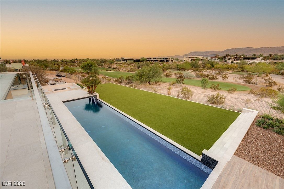 This image showcases a luxurious outdoor space featuring a long, rectangular swimming pool with a tiled edge, adjacent to a well-manicured green lawn. The pool area is bordered by a modern patio with glass railings, offering an elevated view of the surrounding desert landscape, golf course, and distant mountains. The scene evokes a sense of tranquility and upscale living.