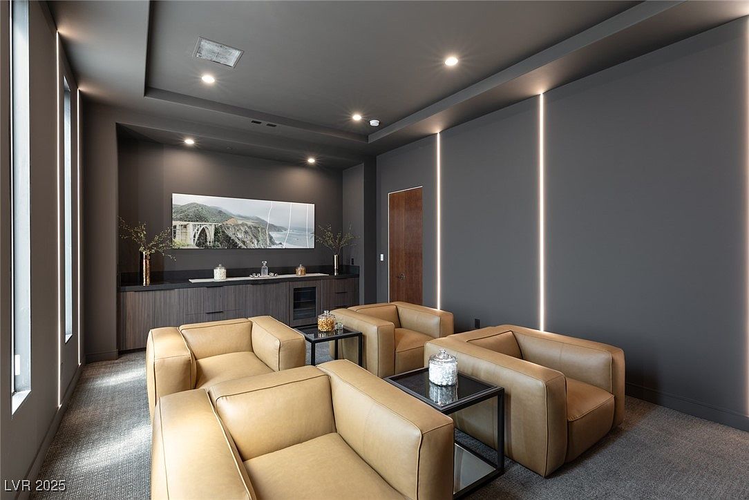 This interior shot showcases a modern living room with a sophisticated color palette of grays and tans. The room features four tan leather armchairs arranged around small black side tables, creating a cozy seating area. A built-in dark wood cabinet with a countertop displays decorative items and a large landscape artwork, while vertical LED light strips add a contemporary touch to the walls.