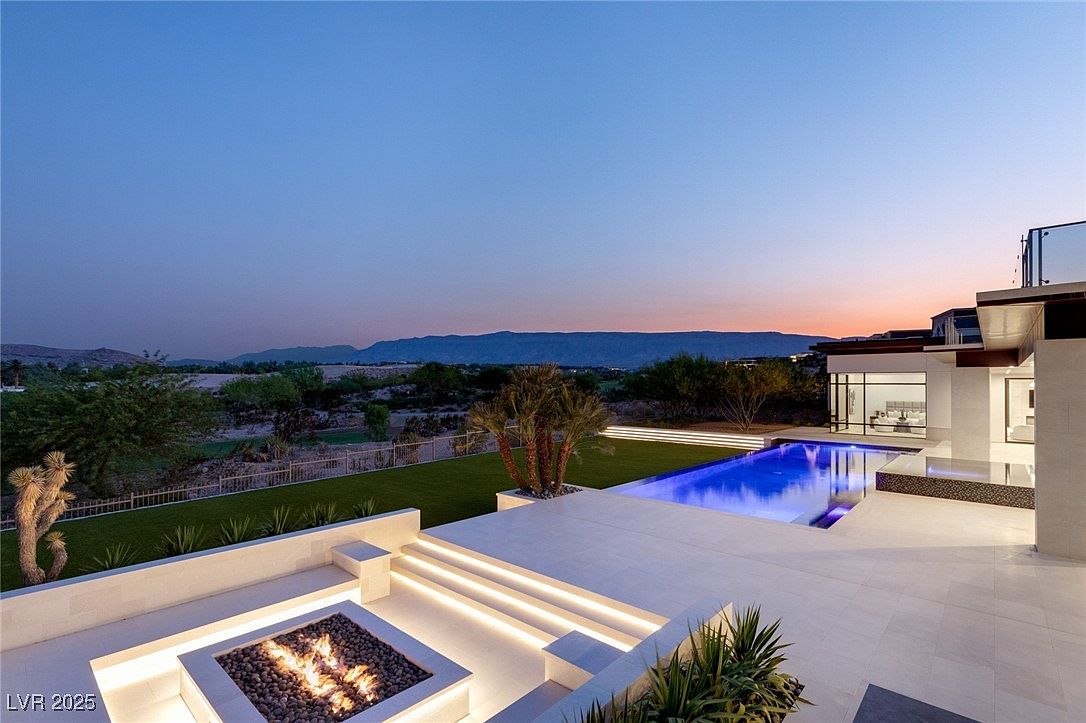 This image showcases a luxurious backyard with a modern aesthetic, featuring a sleek pool illuminated with blue light, a fire pit with glowing flames, and manicured landscaping. The outdoor space includes a contemporary patio with built-in seating and steps, all set against a backdrop of distant mountains and a twilight sky. The overall impression is one of high-end relaxation and sophisticated outdoor living.