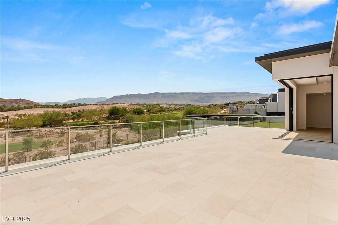 This image showcases a spacious patio or balcony area with a modern design. The flooring is made of large, light-colored tiles, and a glass railing provides an unobstructed view of the surrounding landscape, which includes greenery and distant mountains. The area seamlessly connects to the interior through a large opening, suggesting an indoor-outdoor living concept.