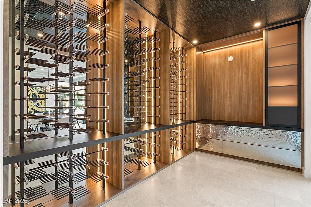 This is a modern wine cellar featuring a unique wine rack system with metal supports and clear acrylic cradles. The walls are paneled with wood and illuminated with vertical LED strips, creating a warm ambiance. A reflective countertop and tiled floor add to the contemporary design, while a cabinet with frosted glass doors provides additional storage.