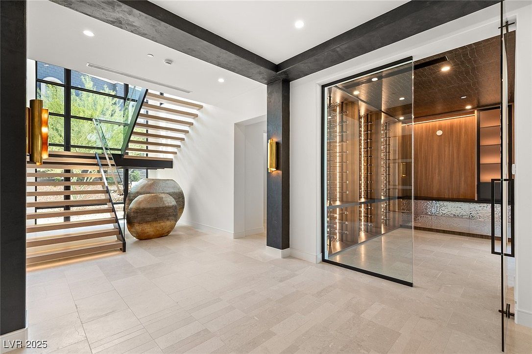 This interior shot showcases a modern hallway with a striking floating staircase and a glass-enclosed wine cellar. The open-concept design features neutral-toned flooring, contemporary lighting fixtures, and large decorative vases. The overall impression is one of luxury and sophisticated design.