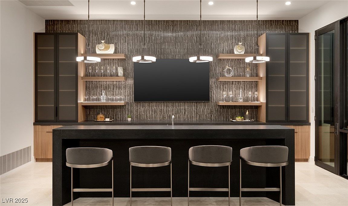 This image showcases a modern wine cellar featuring a dark countertop bar with four gray bar stools. Behind the bar, there's a textured wall with floating shelves displaying decorative items and glassware, flanked by glass-front cabinets. The lighting is provided by modern pendant lights, creating a sophisticated and inviting atmosphere.
