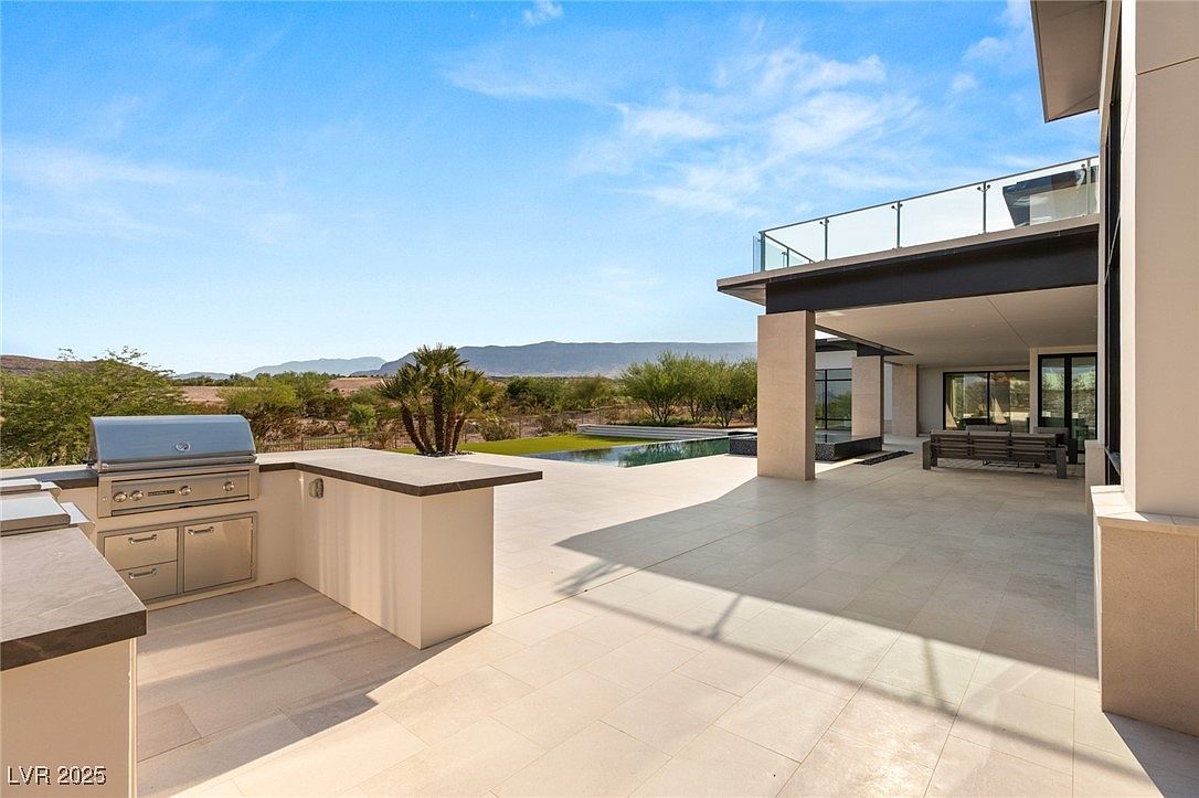 This is a view of a modern home's outdoor patio area, featuring a built-in outdoor kitchen with a stainless steel grill and countertop space. The patio is paved with light-colored tiles, and there's a covered seating area adjacent to the house. In the background, there's a pool, landscaping, and a mountain range, creating a luxurious and inviting outdoor living space.