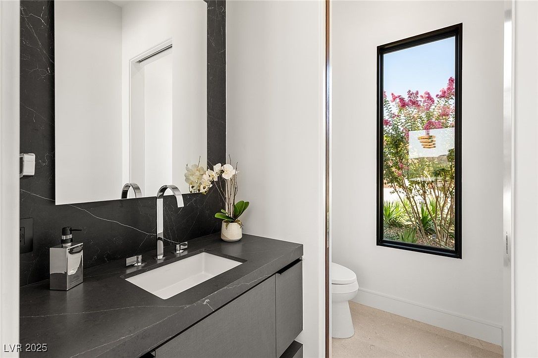 This is a modern bathroom featuring a dark marble countertop and backsplash with a rectangular white sink. A sleek silver faucet and soap dispenser add to the contemporary design. A window provides natural light and a view of the outdoors, while a toilet is visible in the background, suggesting a functional and stylish space.