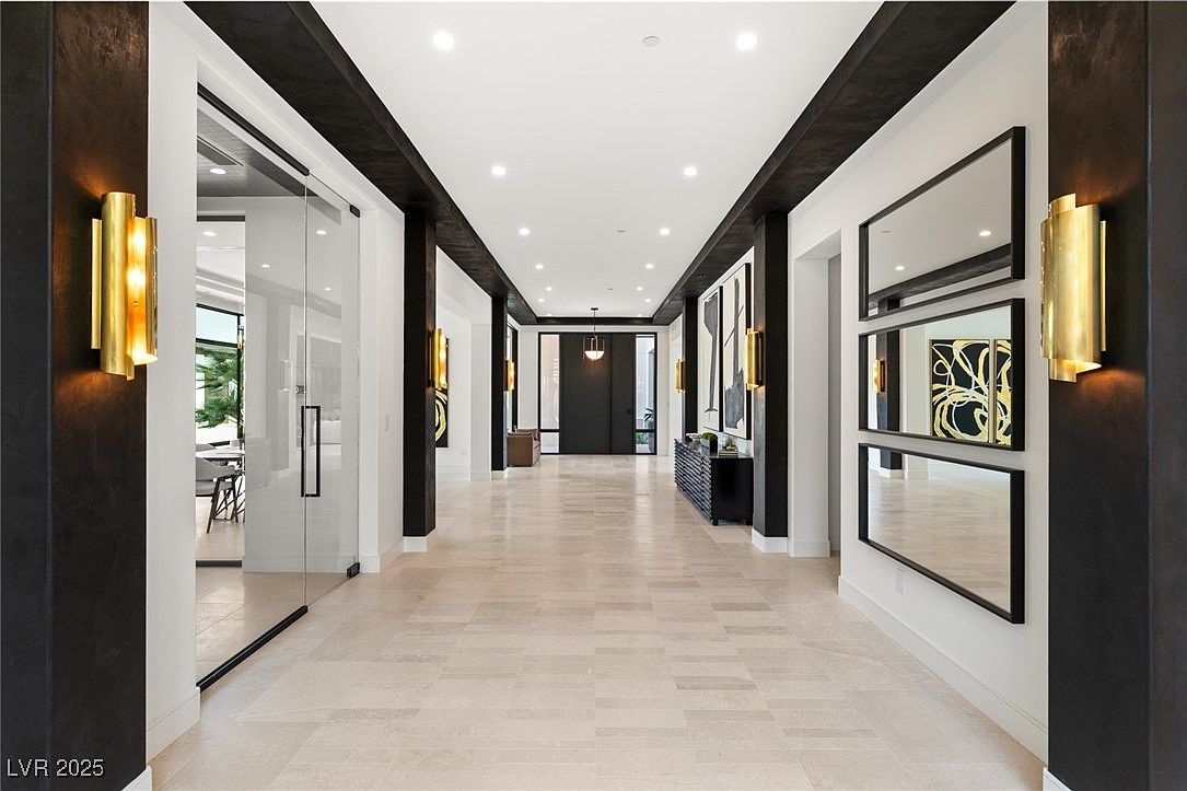 This is an interior shot of a modern hallway featuring dark wood beams and pillars contrasting with white walls and light tile flooring. The hallway is illuminated by recessed lighting and gold sconces, and it includes decorative mirrors and artwork. The perspective is a straight-on view, emphasizing the length and depth of the hallway, creating a sense of spaciousness and luxury.