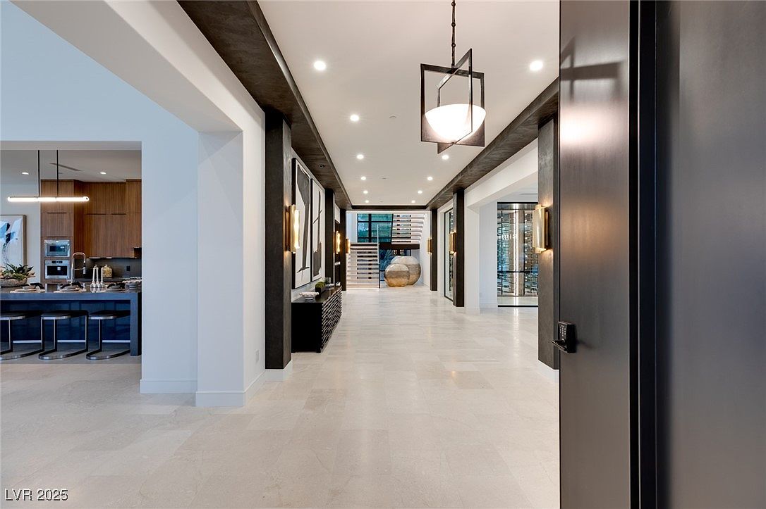This interior shot showcases a modern hallway with a neutral color palette and sophisticated design elements. The hallway features recessed lighting, decorative wall sconces, and a unique pendant light fixture. The flooring is a light-colored tile, and the space leads to a glimpse of a kitchen and a wine cellar, creating an inviting and luxurious atmosphere.