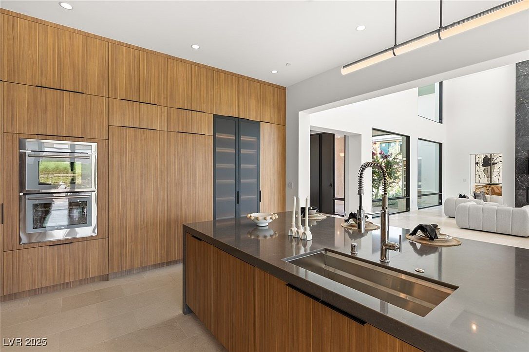 This is a modern kitchen featuring sleek, wood-paneled cabinetry and a dark countertop island with a built-in sink. Stainless steel appliances are integrated into the cabinetry, and a contemporary light fixture hangs above. The open layout leads into a living area, creating a spacious and inviting atmosphere.