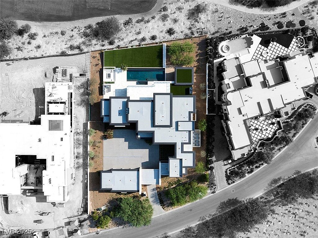 This aerial view showcases a modern luxury home with a geometric roof design, a rectangular swimming pool, and manicured green spaces. The property is surrounded by desert landscaping and neighboring estates, highlighting its exclusivity and privacy. A large driveway and multiple structures suggest ample space and functionality.