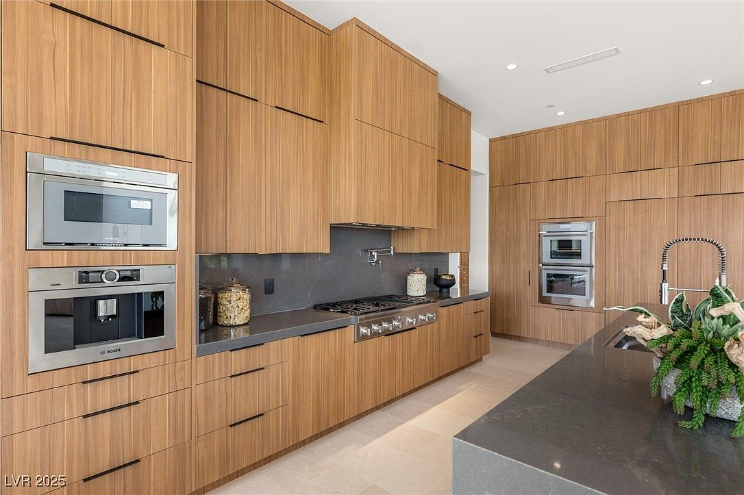 This is a modern kitchen featuring sleek, wood-paneled cabinetry with minimalist hardware. Stainless steel appliances, including a built-in microwave, oven, and coffee maker, are seamlessly integrated into the design. The countertops are a dark gray, providing a sophisticated contrast to the wood, and the overall impression is one of clean lines and contemporary elegance.