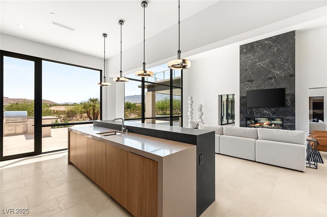 This is a bright, modern kitchen featuring a large island with a light countertop and wood cabinetry. The kitchen opens to a living area with a fireplace and a large window offering an outdoor view. The space is well-lit with pendant lighting and natural light, creating a clean and luxurious atmosphere.