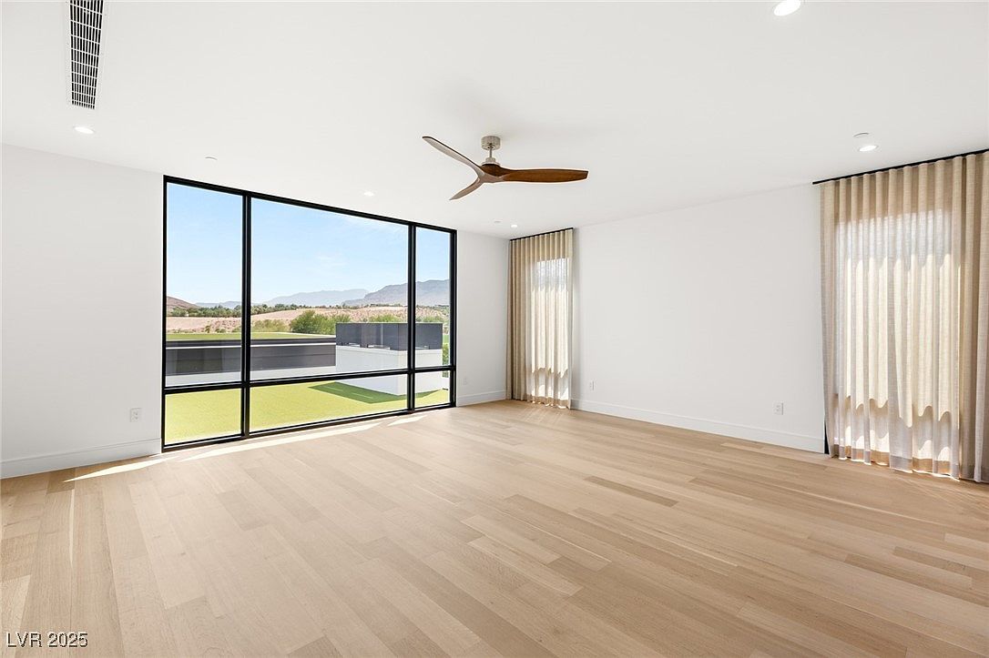 This is a bright and spacious primary bedroom featuring large windows that offer an expansive view of the landscape. The room has light hardwood floors, white walls, and a modern ceiling fan. A neutral-toned curtain adds a touch of elegance to the space, creating a serene and inviting atmosphere.