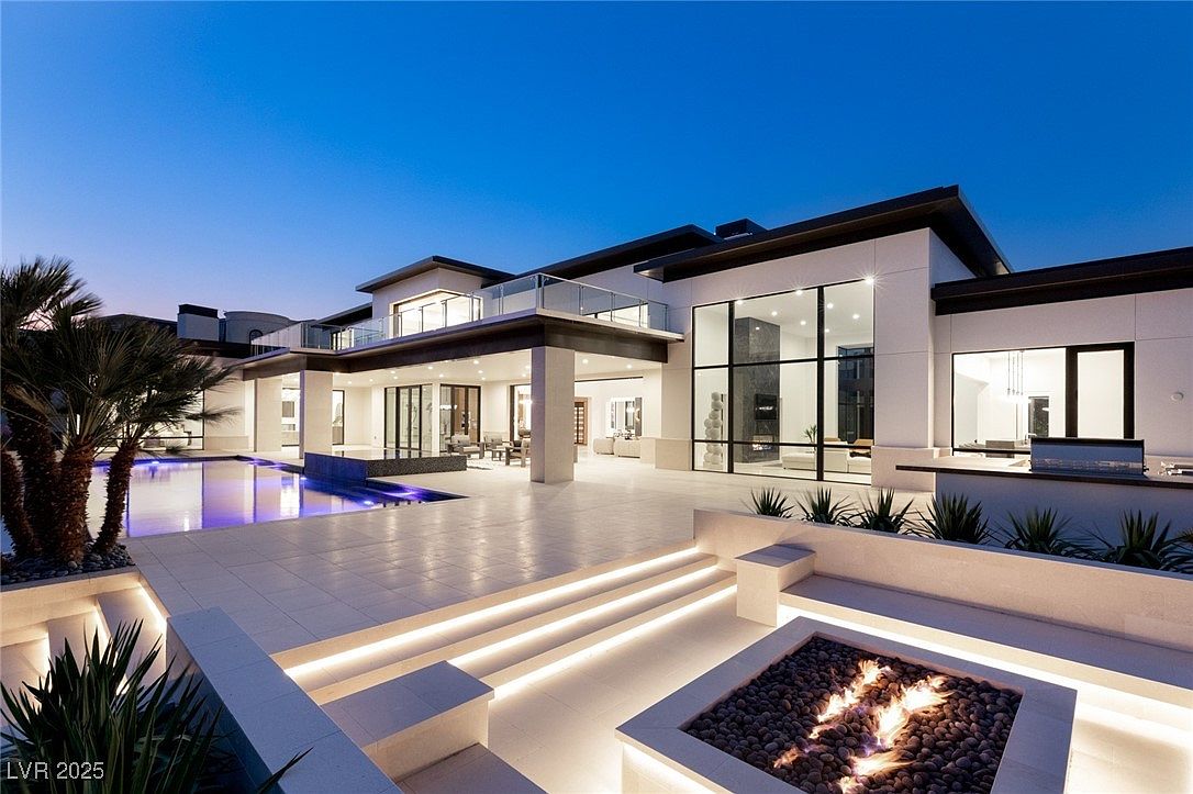 This is a stunning rear view of a modern luxury home at dusk, showcasing a seamless blend of indoor and outdoor living. The property features a large pool with integrated water features, a fire pit area with built-in seating, and expansive patio space perfect for entertaining. Large windows and sliding glass doors provide ample natural light and views into the home's interior, while the architectural design emphasizes clean lines and sophisticated elegance.