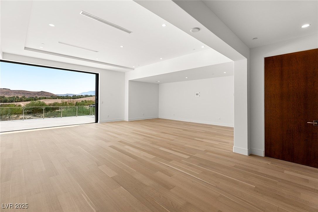 This is a bright and spacious living room with light hardwood floors and white walls. A large sliding glass door provides access to an outdoor balcony and offers a scenic view of the surrounding landscape. A dark wood door is visible on the right side of the room, adding a touch of contrast to the otherwise light color palette.
