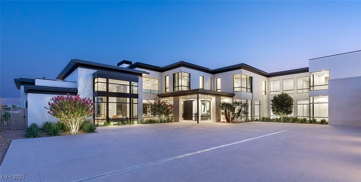 This is a front exterior view of a modern, luxurious home. The house features a flat roof, large windows with black frames, and a neutral-colored facade. A spacious driveway leads up to the entrance, and manicured landscaping adds to the property's curb appeal.